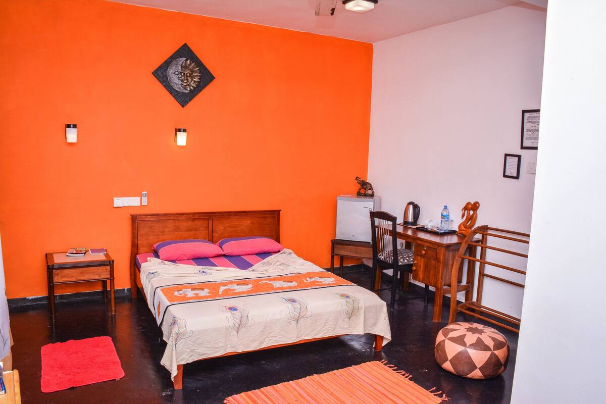 A room with an orange wall, a wooden bed with multiple pillows, a desk with a chair, and a round rug.