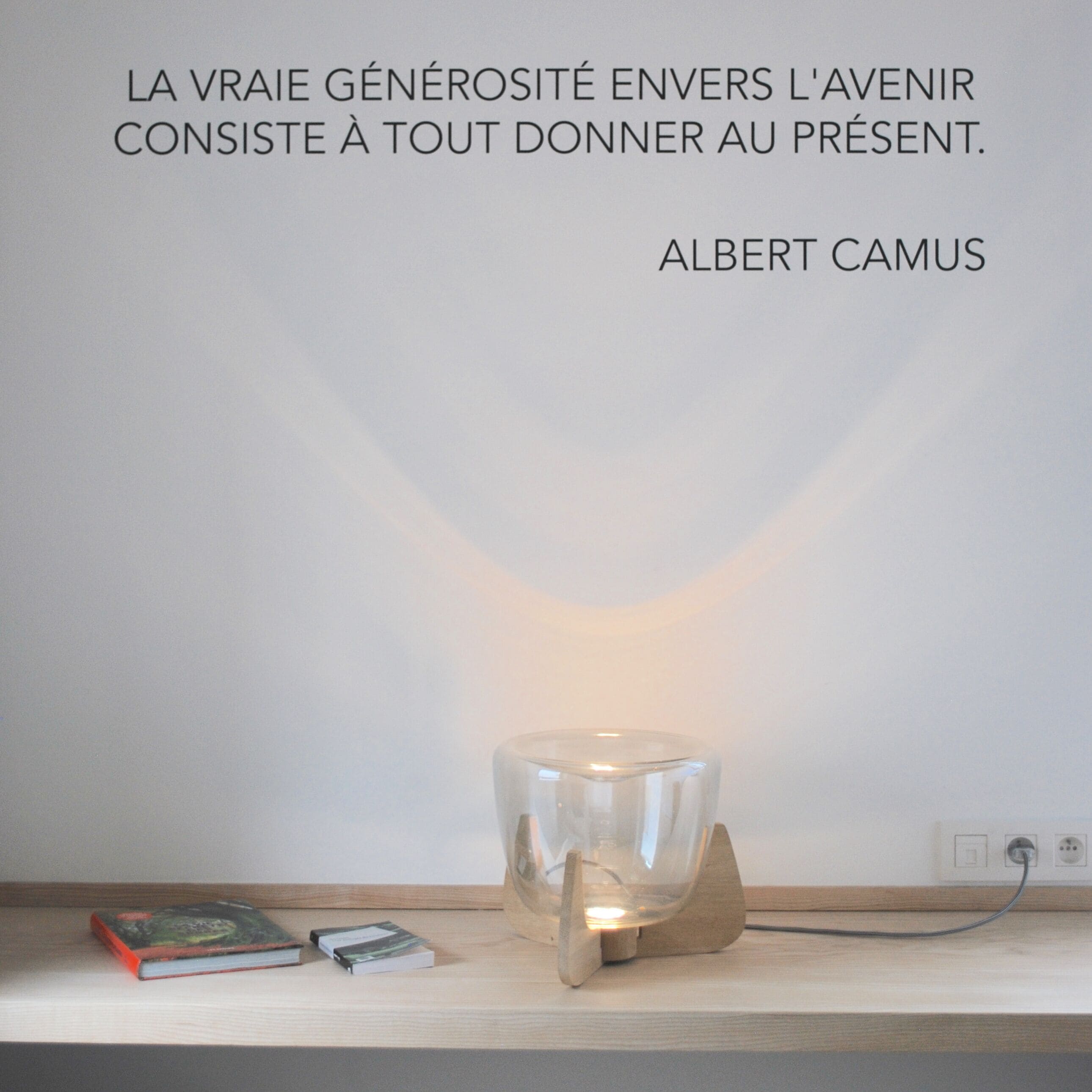 Una pared con una cita de Albert Camus y una mesa con libros y una lámpara de vidrio.