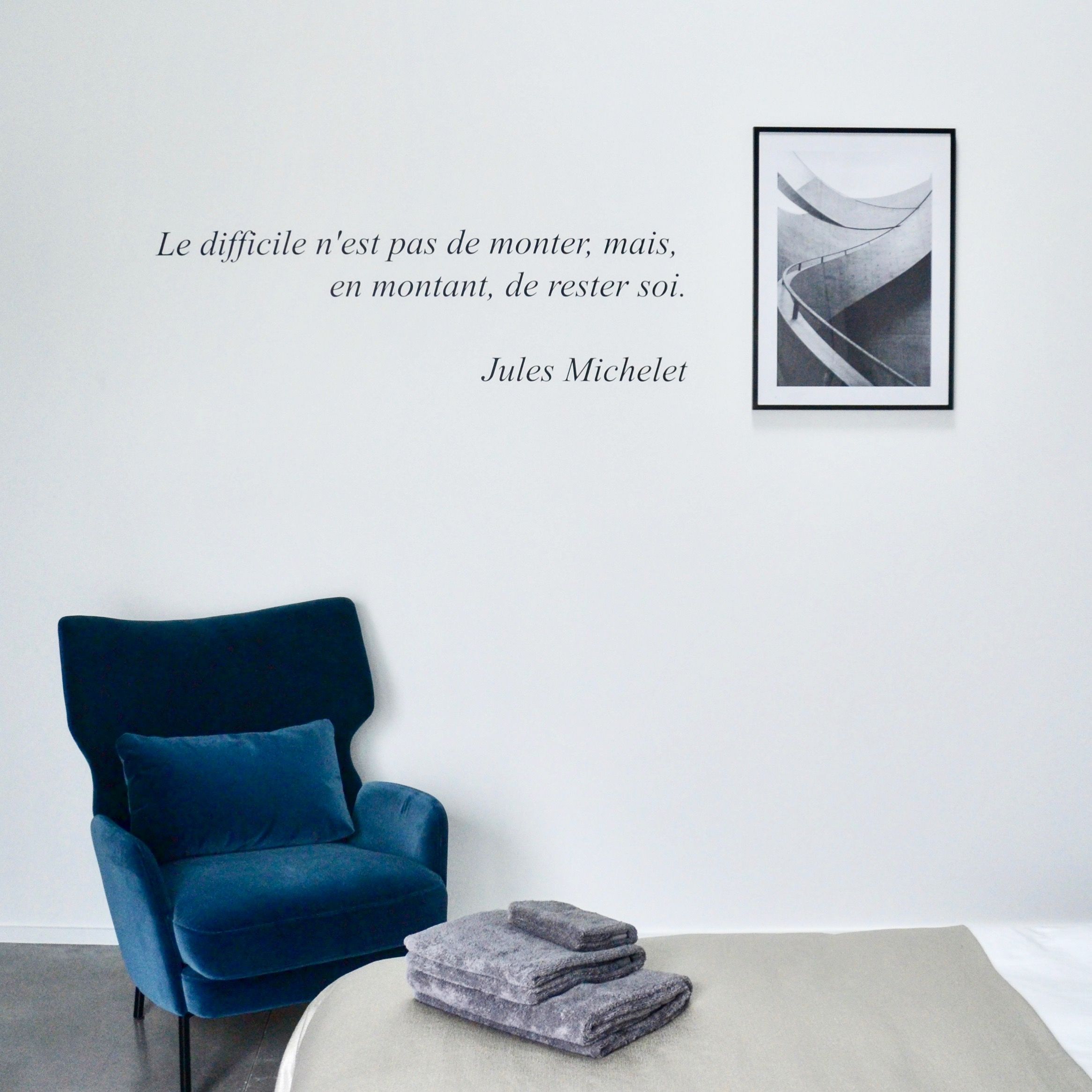 A modern room with a blue armchair, a framed quote on the wall, and a small table with folded towels.