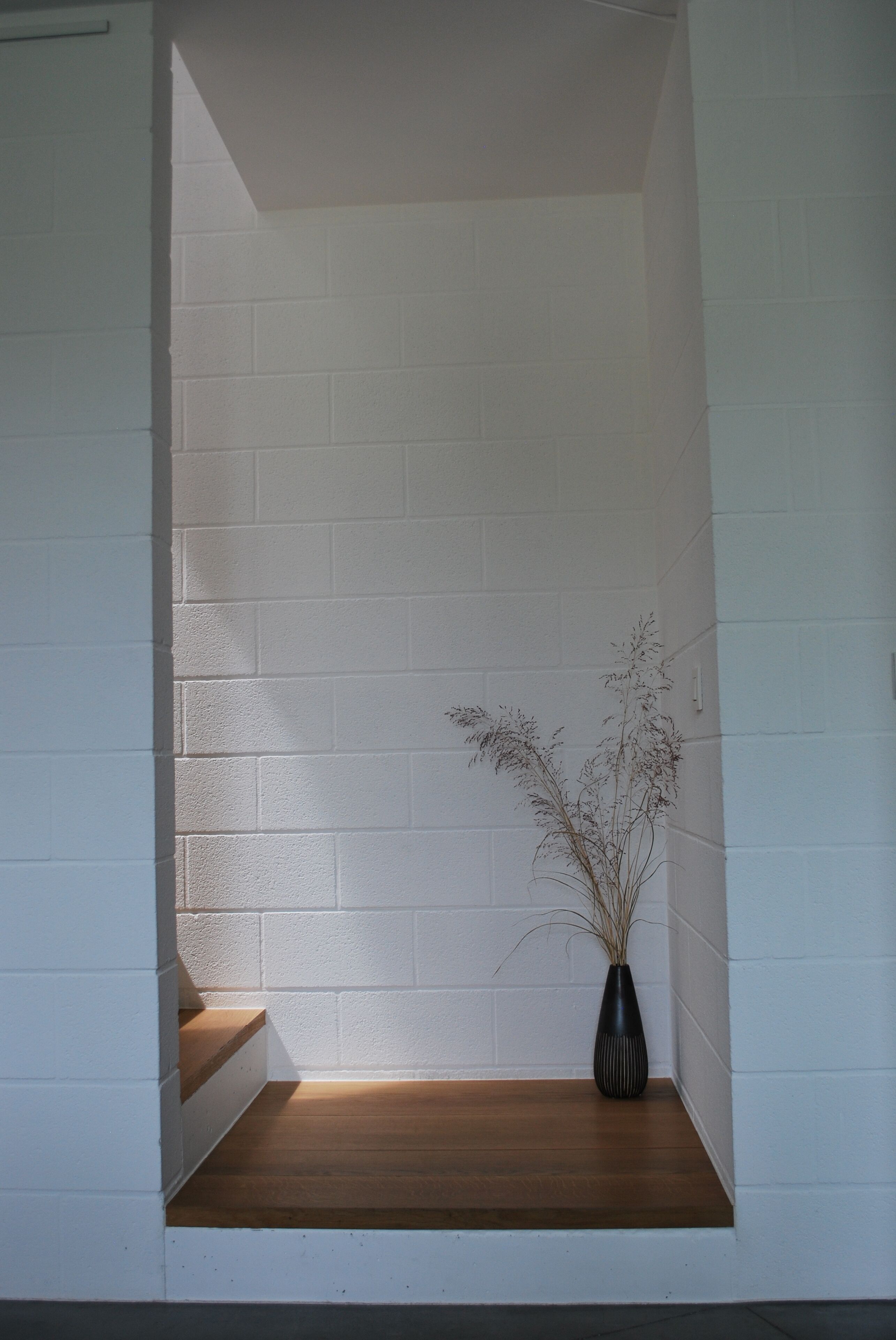 A corner with white brick walls, a wooden bench, and a black vase with dried plants.