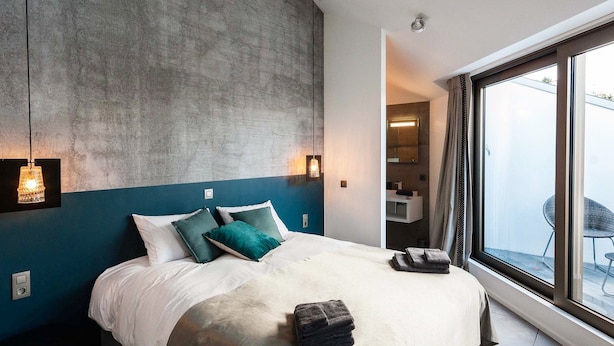 A modern bedroom with a large bed, a concrete accent wall, and a view of a balcony. - Dimensions M (Mont-Saint-Guibert)