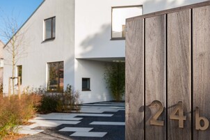 Un edificio moderno de diseño minimalista, con una cerca de madera que muestra el número '24b' y un sendero blanco y negro que lleva a la entrada.