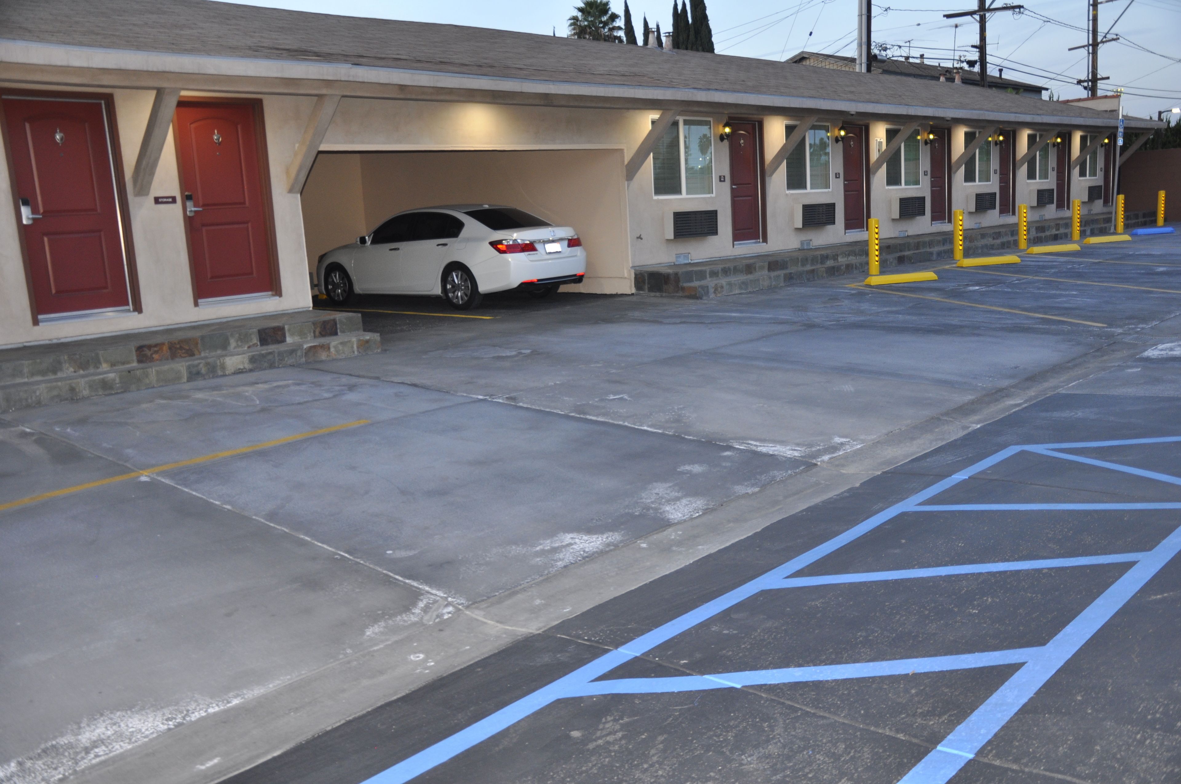 Un motel con un coche blanco estacionado en el garaje, una fila de habitaciones con puertas rojas y un estacionamiento con espacios reservados para discapacitados azules.