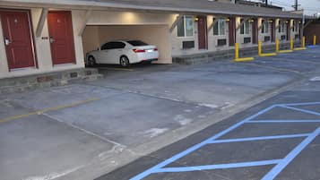 Un motel con un coche blanco estacionado en el garaje, una fila de habitaciones con puertas rojas y un estacionamiento con espacios reservados para discapacitados azules.
