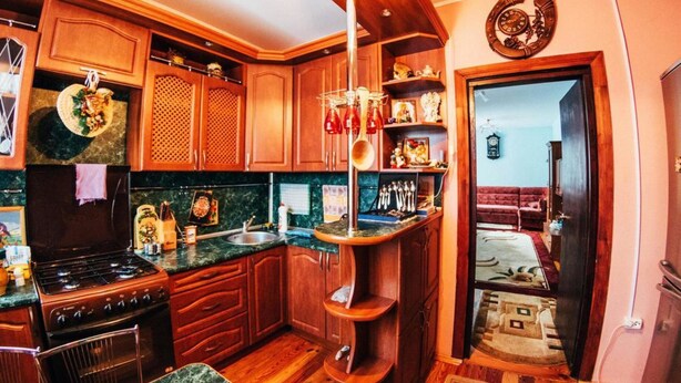 Une cuisine avec des armoires en bois, une cuisinière et un évier. On y trouve une étagère en bois sur laquelle sont disposés divers objets, ainsi qu’une horloge au mur.