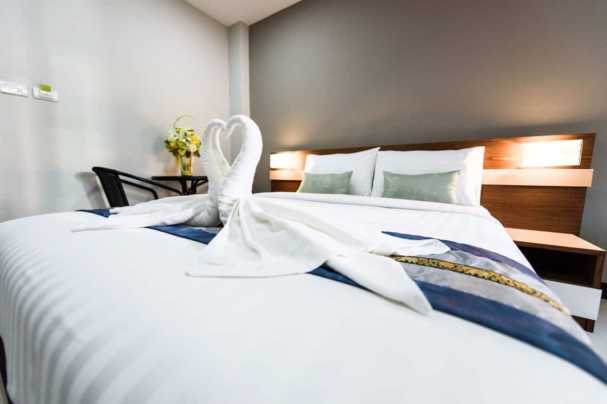 A neatly made bed with a heart-shaped towel arrangement, a bedside table with a lamp, and a wall-mounted light fixture.