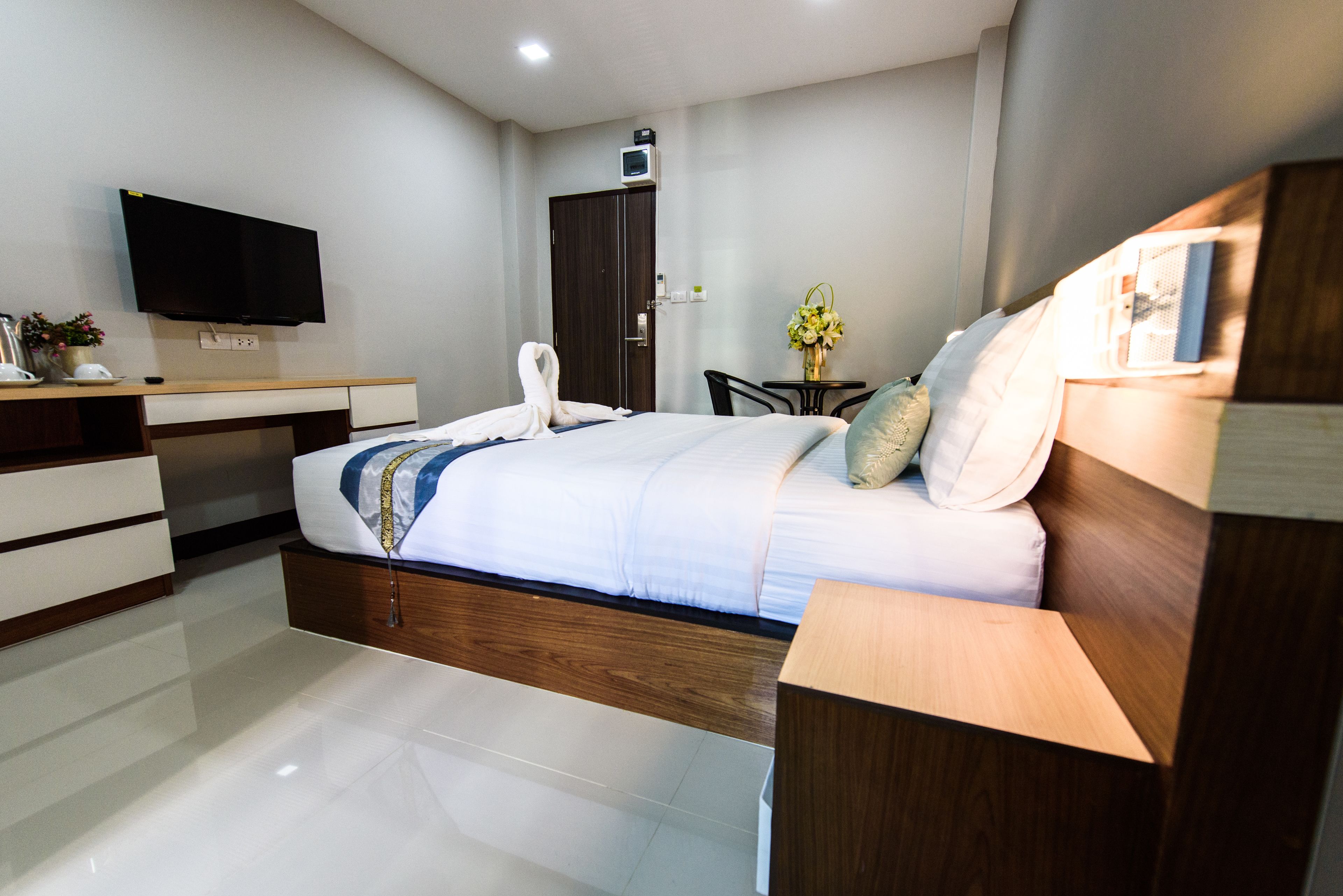 Un dormitorio moderno con cama, cabecera de madera, mesita de noche, televisor, escritorio, silla y una pequeña mesa con flores.