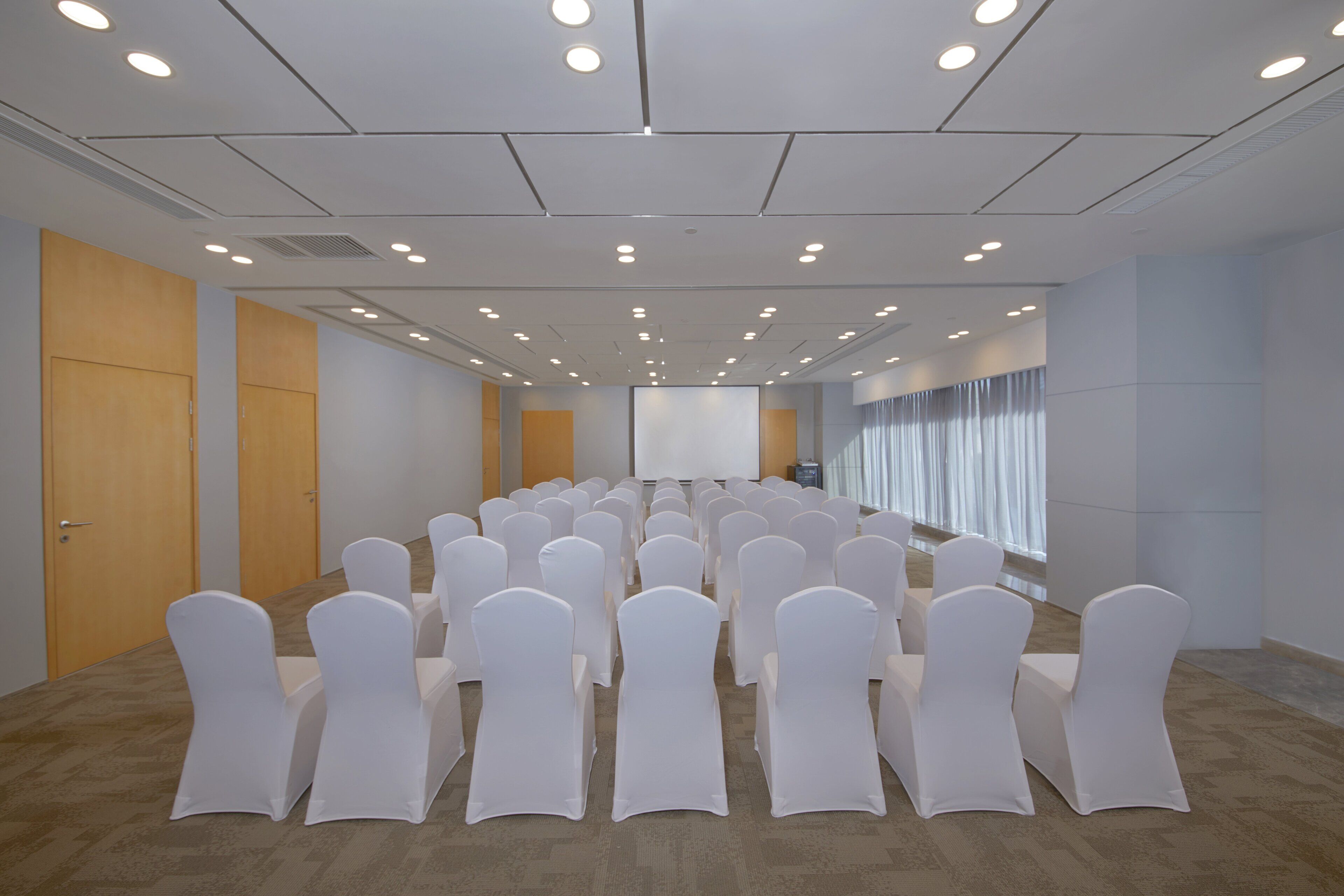 Une salle de conférence avec des rangées de chaises blanches, un écran de projection et une grande fenêtre avec des rideaux.