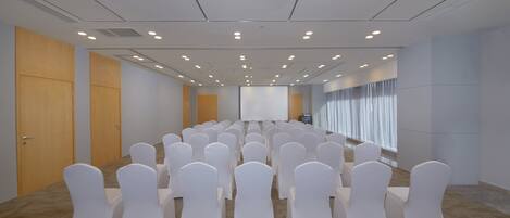 Une salle de conférence avec des rangées de chaises blanches, un écran de projection et une grande fenêtre avec des rideaux.