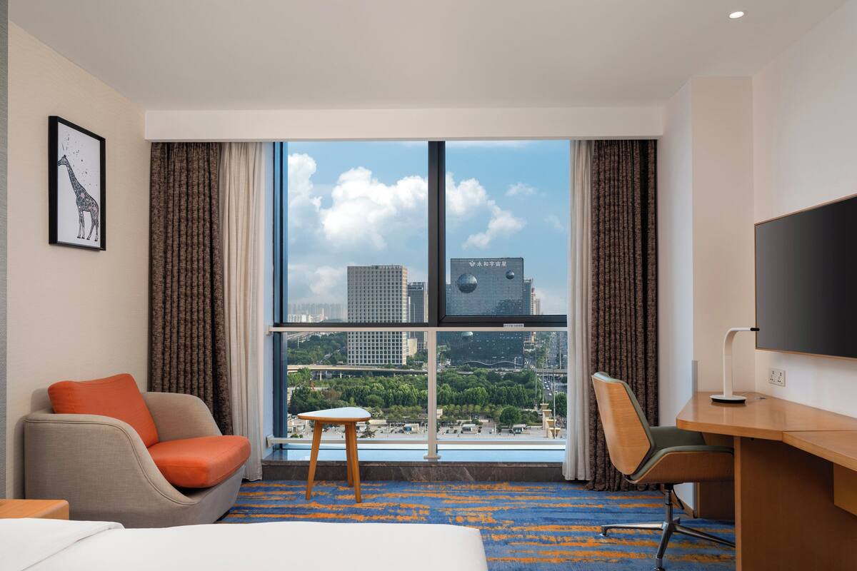 Ein Hotelzimmer mit Bett, Schreibtisch, Stuhl, einem kleinen Tisch und einem großen Fenster mit Blick auf die Stadt.