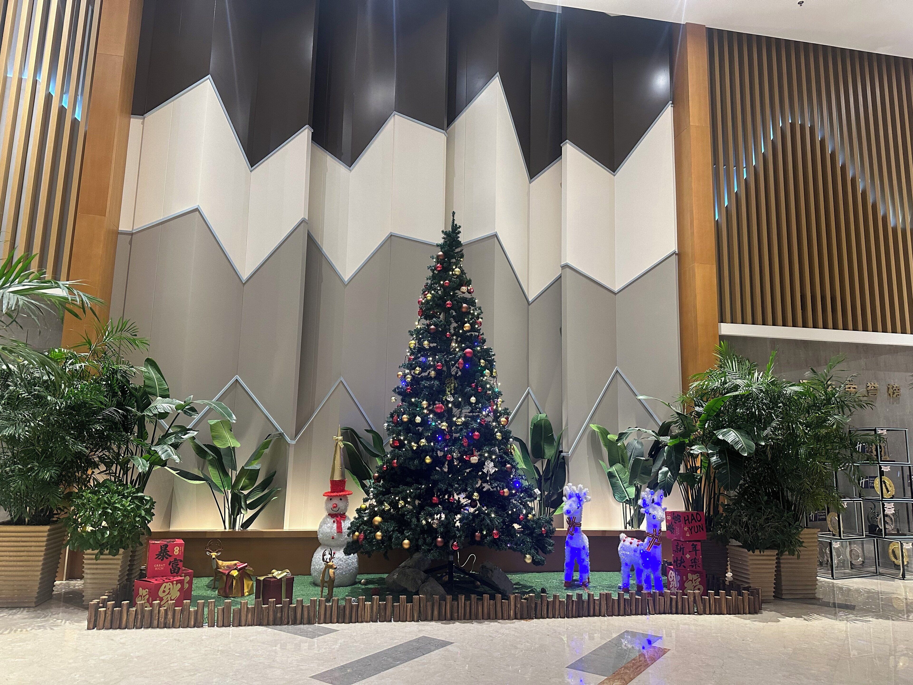 A decorated Christmas tree in a lobby with a geometric wall design and potted plants.