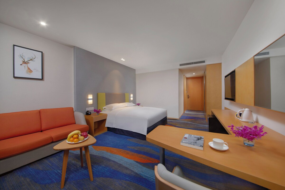 Une chambre d’hôtel avec un lit, un canapé orange, une petite table avec des fruits, un bureau avec une tasse et des fleurs, et une télévision.