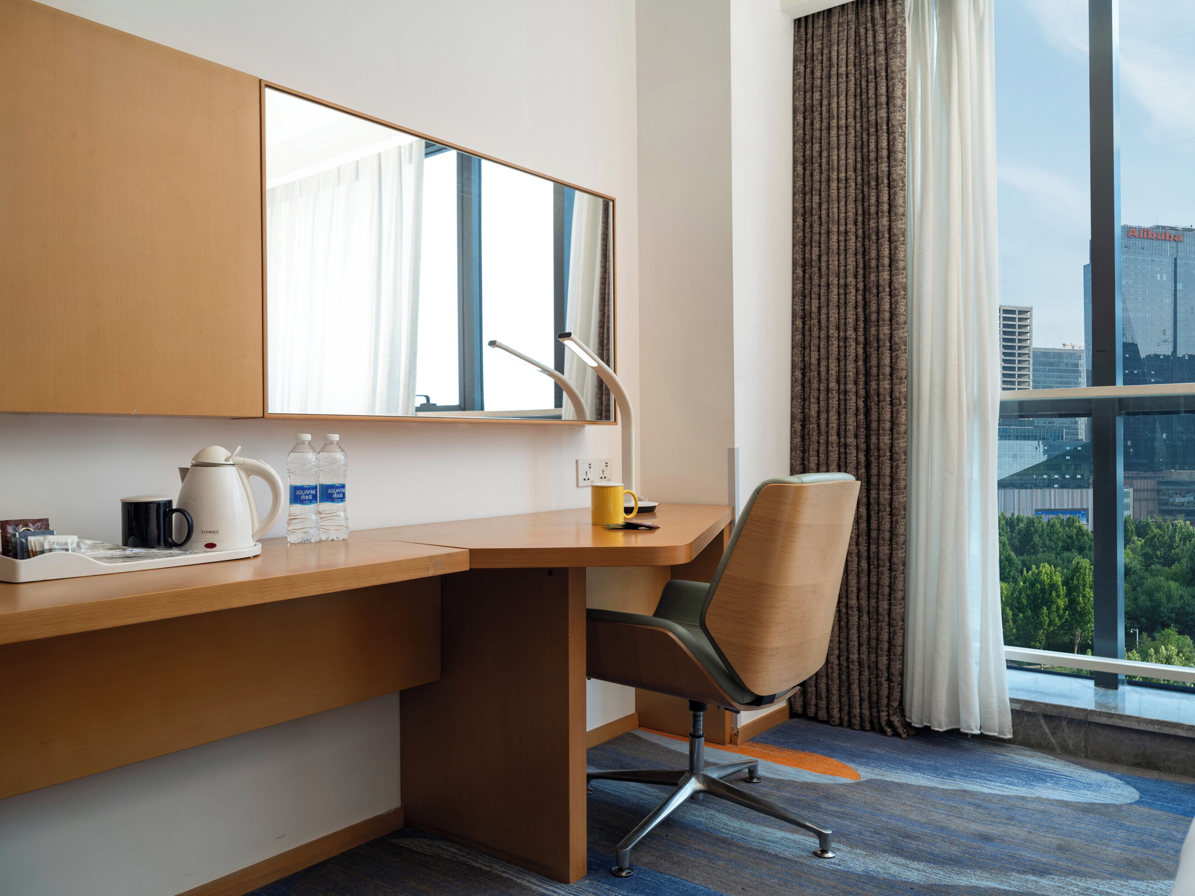 Habitación de hotel con un escritorio de madera, una silla, un espejo, una ventana con cortinas y vistas a un paisaje urbano.
