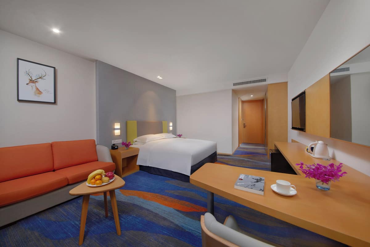 Ein Hotelzimmer mit Bett, orangefarbenem Sofa, kleinem Tisch mit Früchten, Schreibtisch mit Tasse und Blumen sowie einem Fernseher.