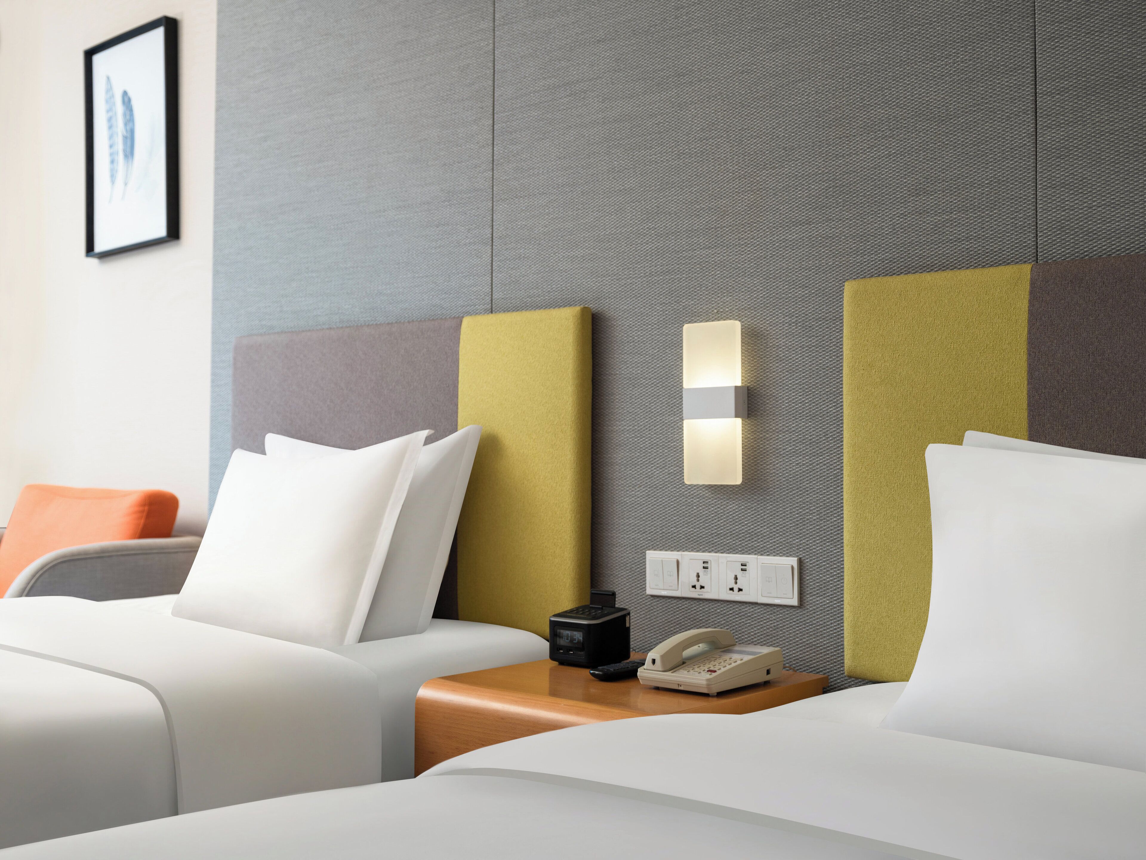 A hotel room with two beds, a bedside table with a telephone and a remote control, and a wall-mounted light fixture.