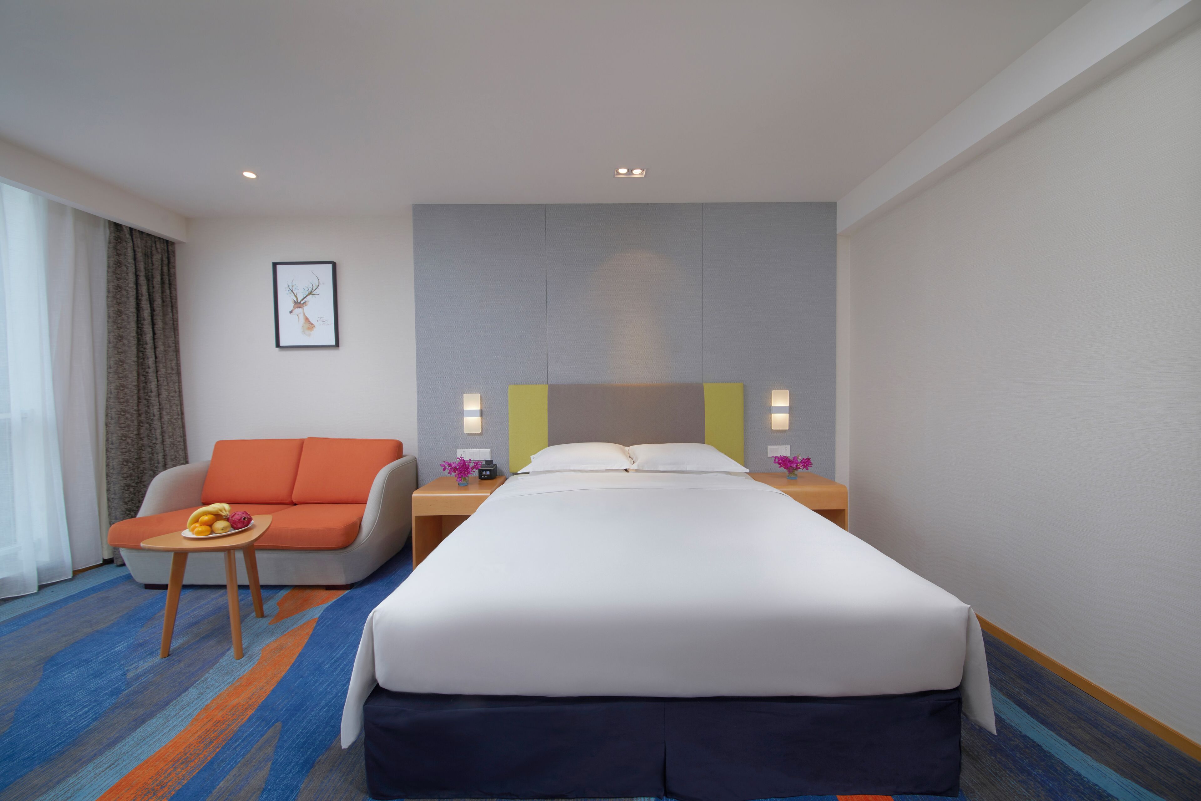 Habitación de hotel con cama, sofá, una mesita con frutero y un cuadro en la pared.