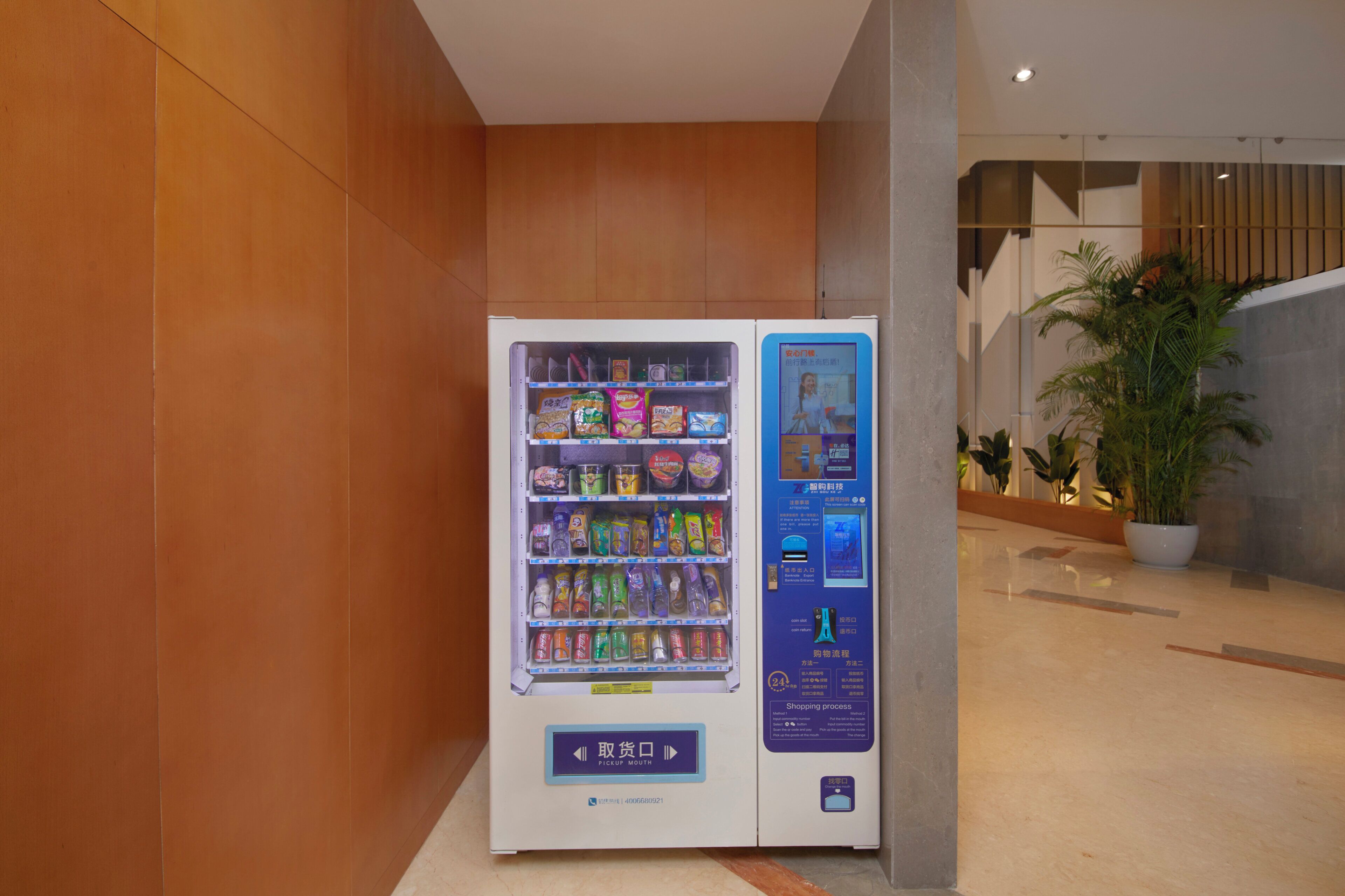 Een automaat met diverse snacks en drankjes, gelegen in een gang met houten wanden en een potplant.