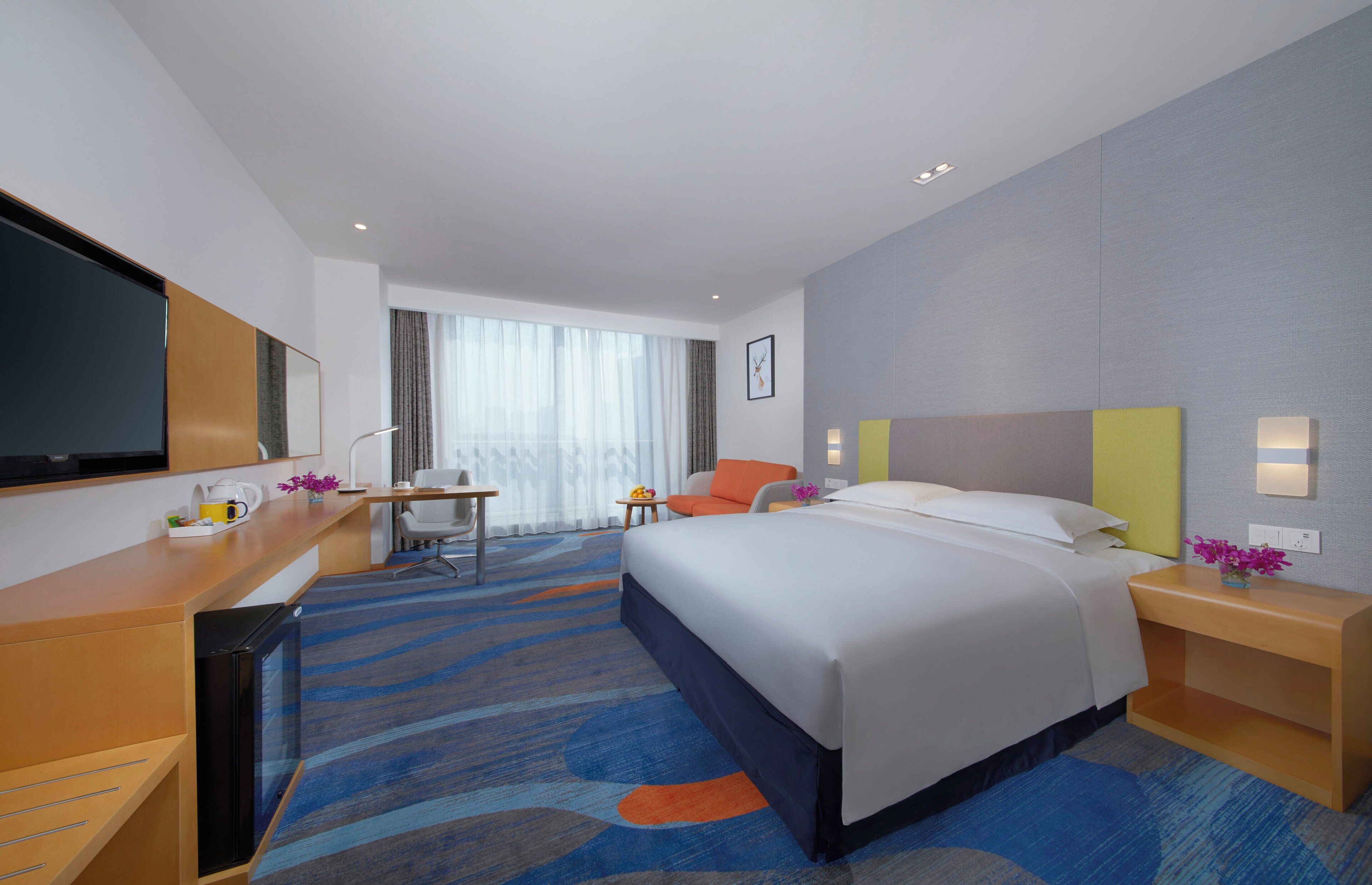 A hotel room with a bed, a desk, a chair, a TV, and a window with curtains.