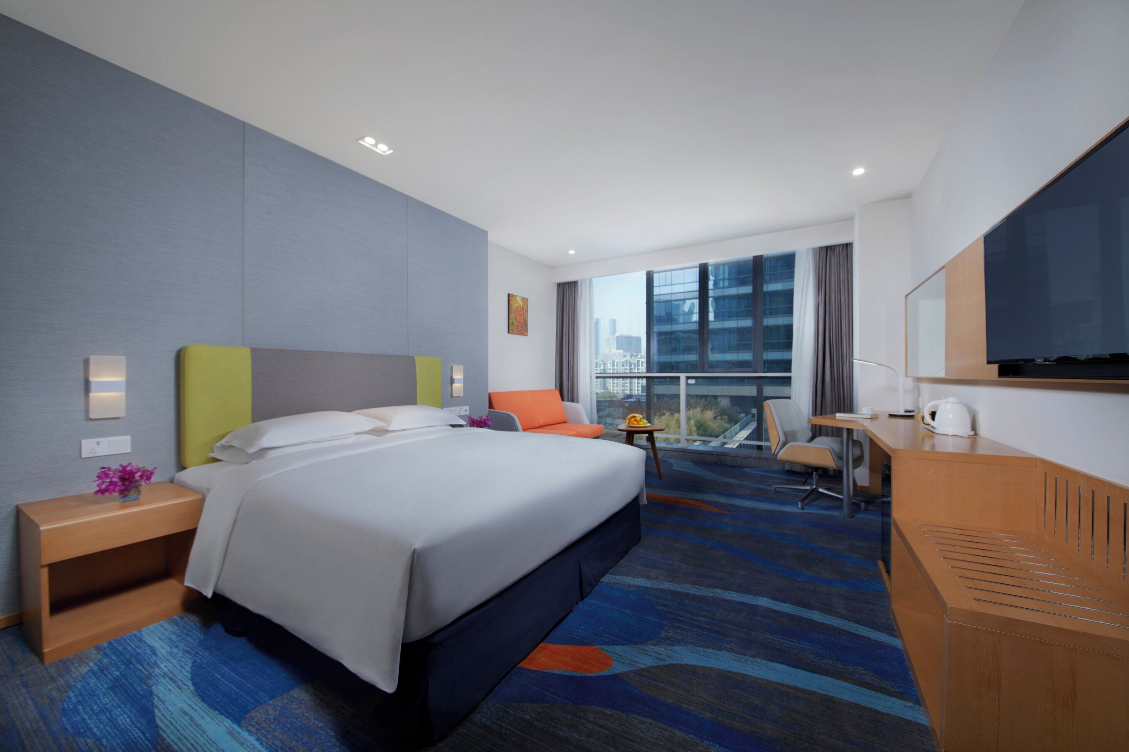 A hotel room with a bed, a desk, a chair, a nightstand, a TV, and a window with a city view.