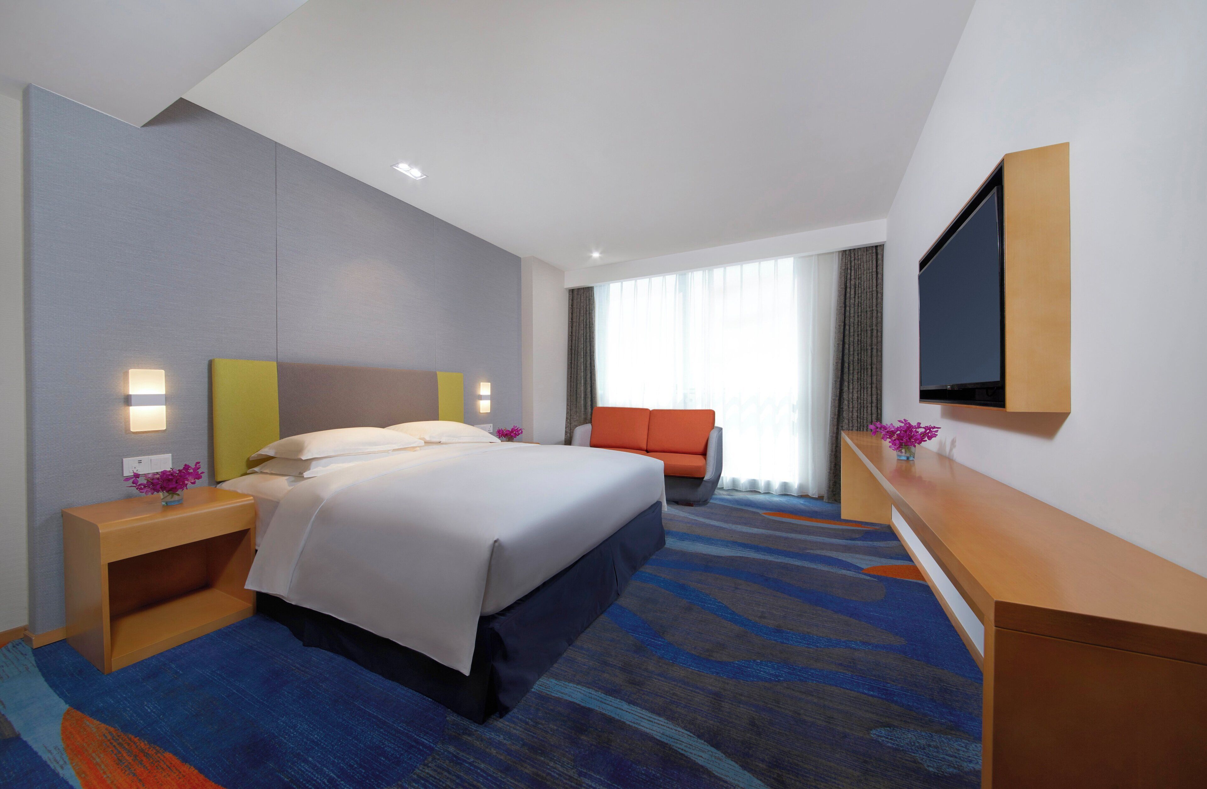 A modern hotel room with a large bed, a flat-screen TV, a desk, and a comfortable seating area.