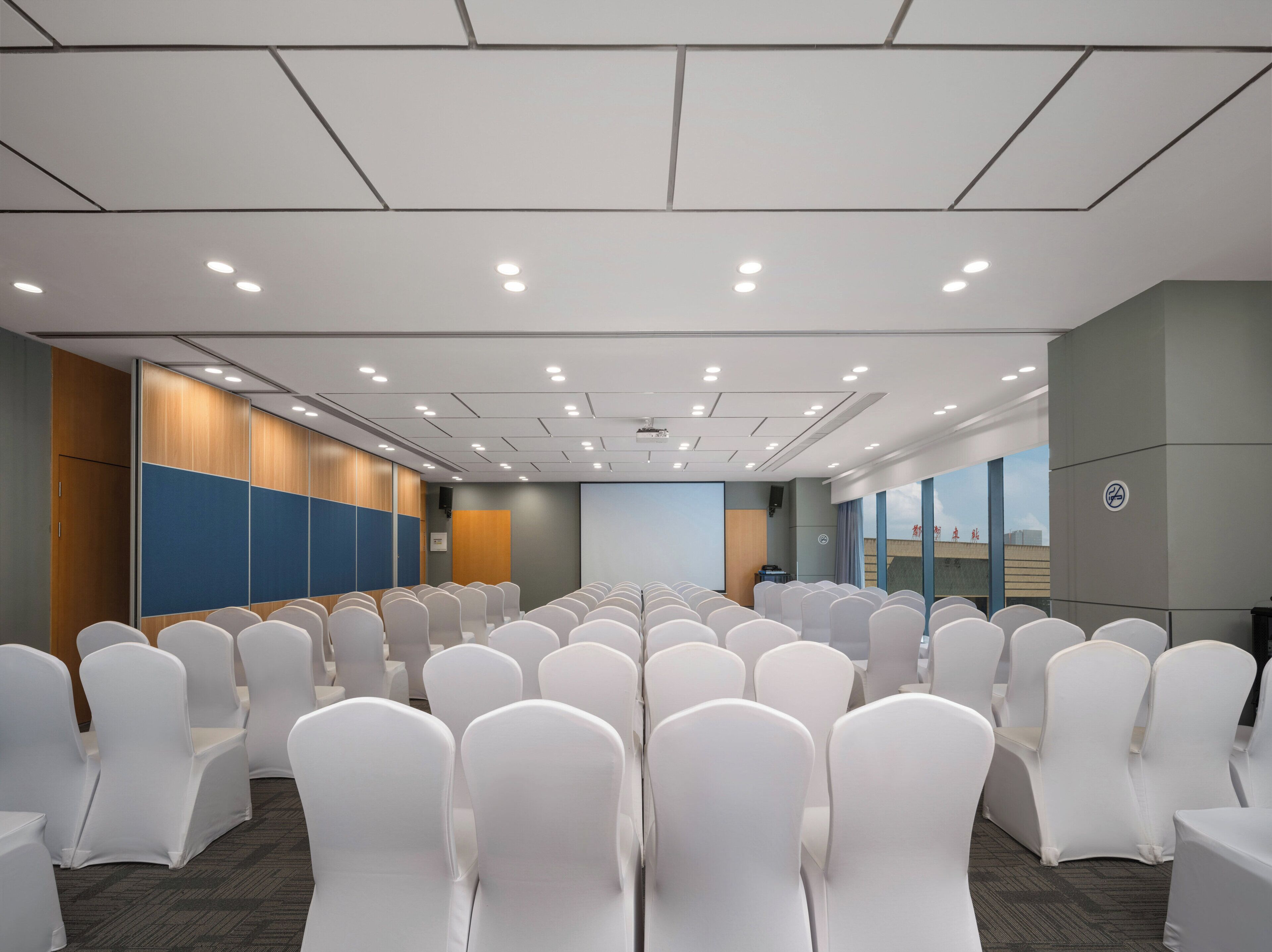 A conference room with rows of white chairs, a projector screen, and a large window with a cityscape view.