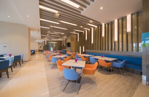 A modern dining area with multiple tables and chairs, a long bench, and a high ceiling with linear lighting.