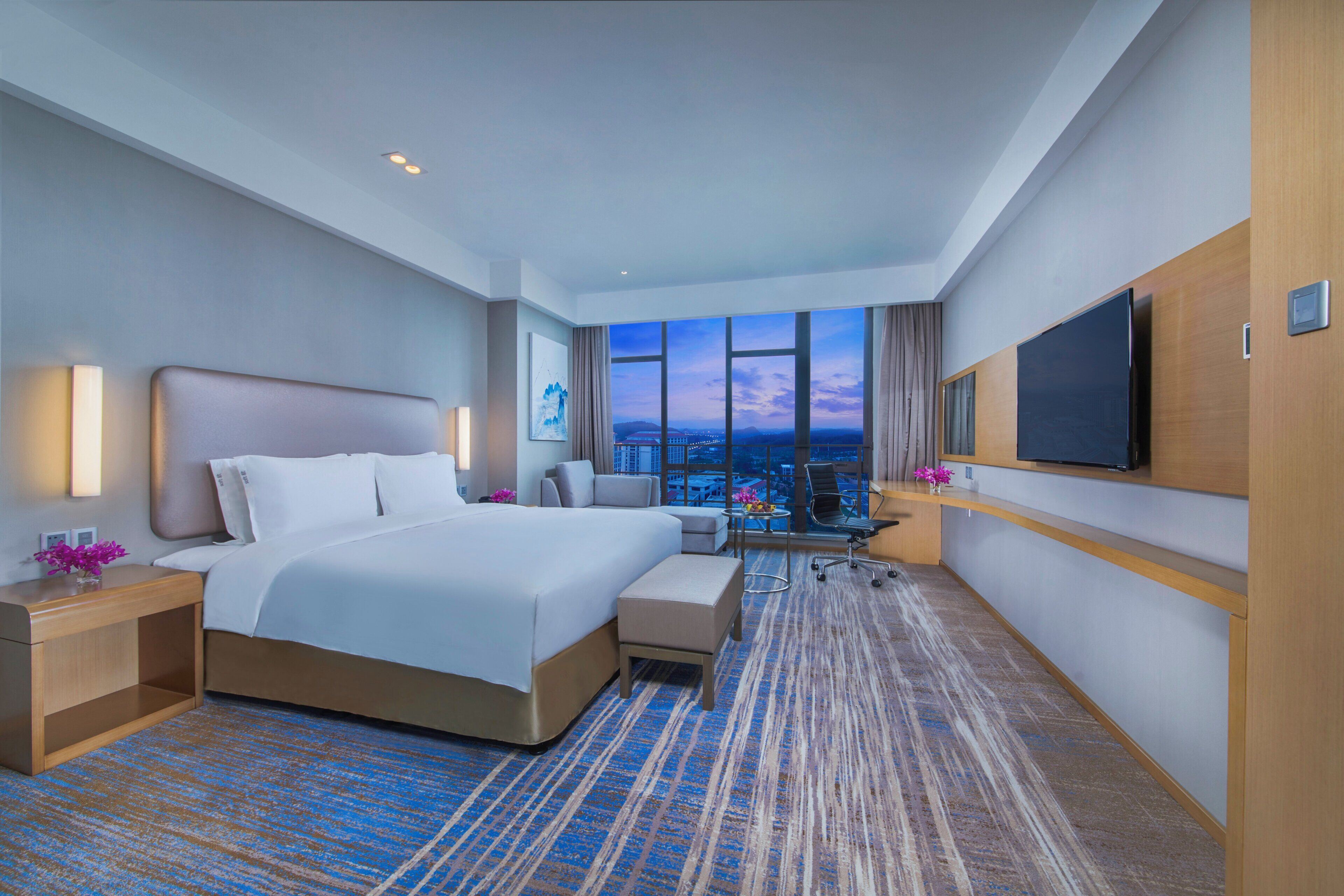 A hotel room with a large bed, a desk, a chair, a TV, and a view of the cityscape.