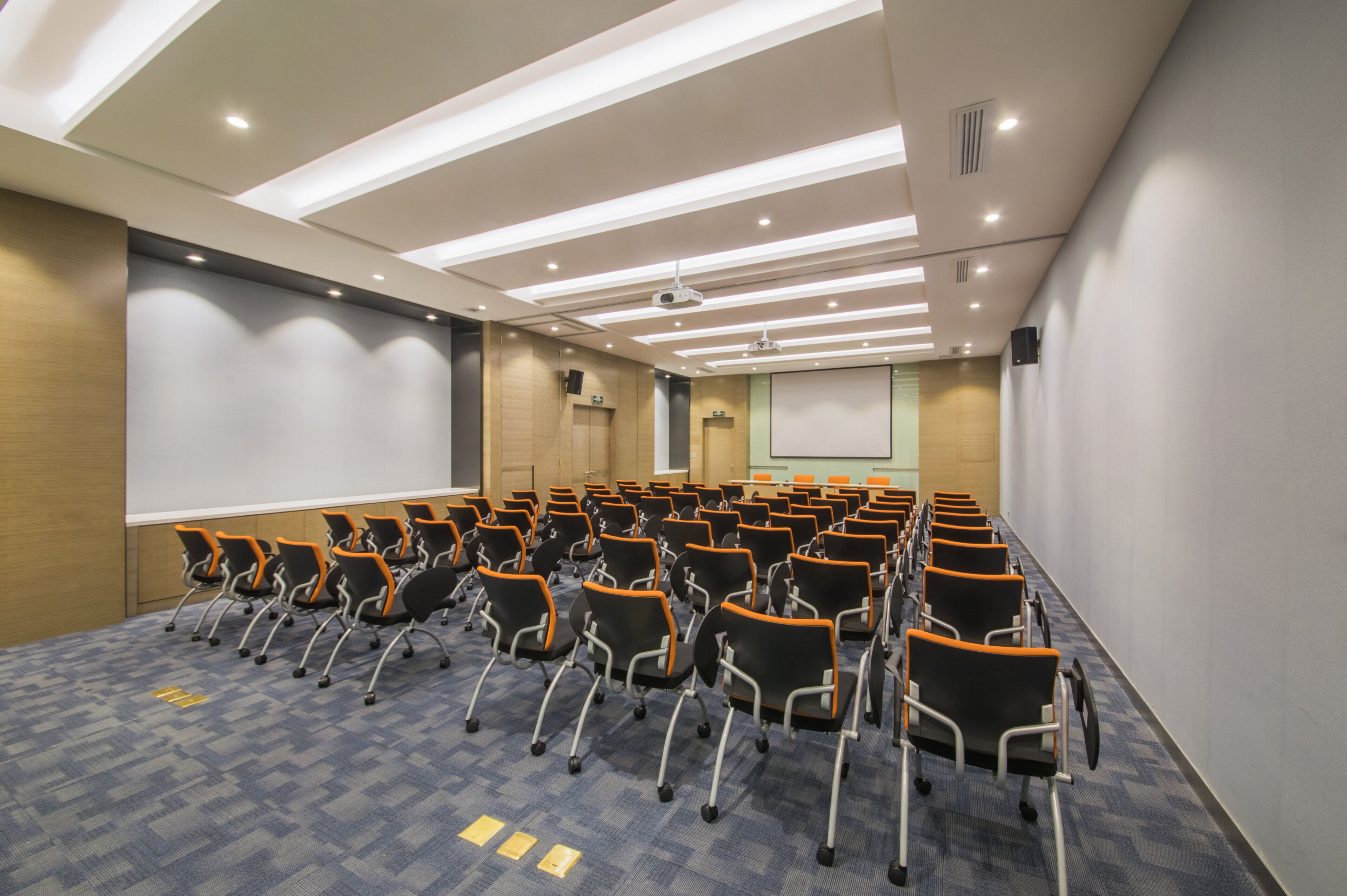 Un salón de conferencias con filas de sillas, una pantalla de proyector y una pantalla para presentaciones.