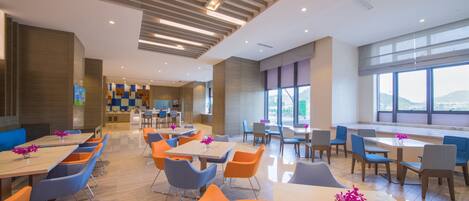 A modern dining area with multiple tables and chairs, a kitchen in the background, and large windows offering a view.