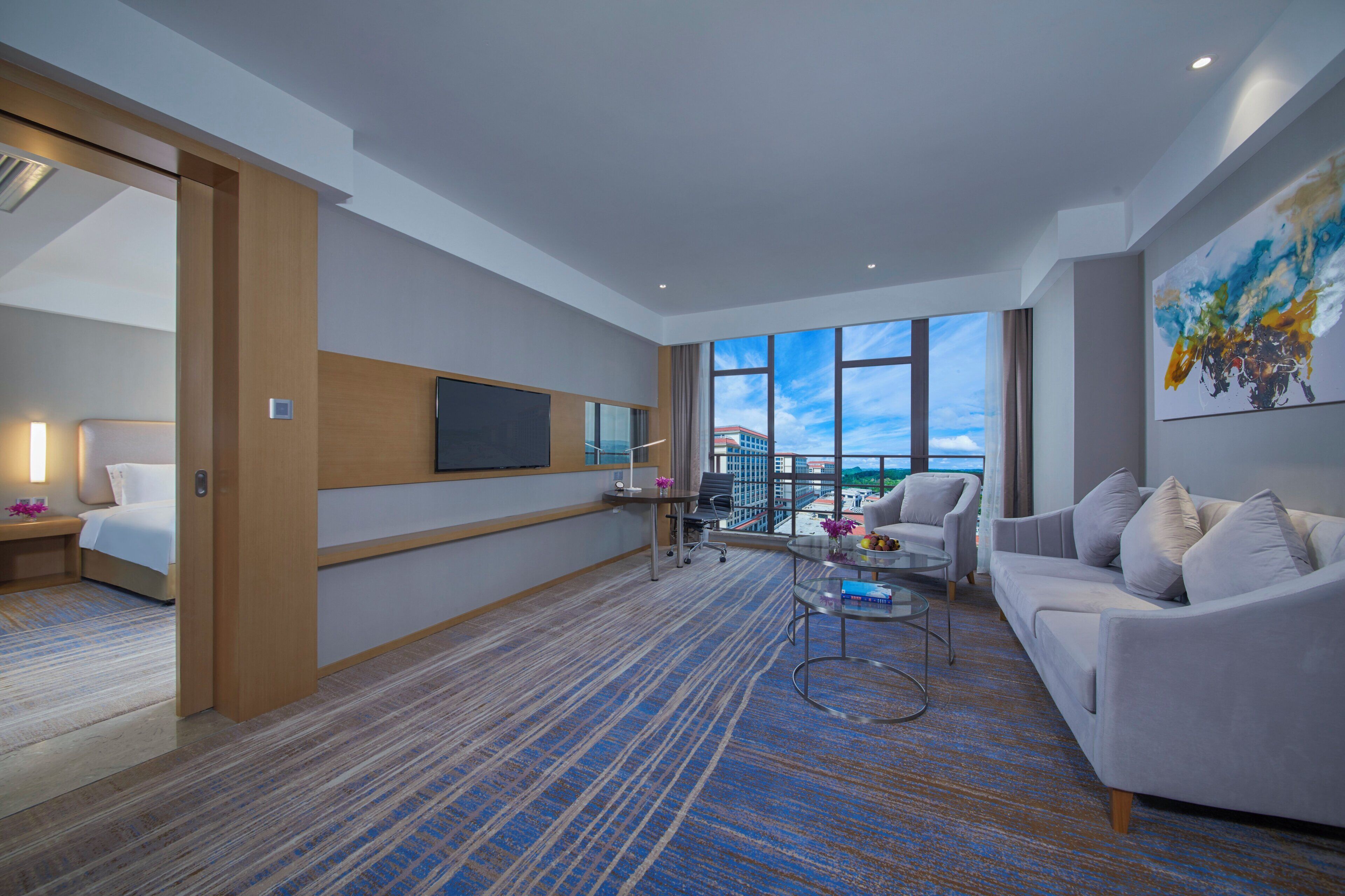 Een moderne hotelkamer met een groot raam, een flatscreen televisie, een bank, een bijzettafeltje en een schilderij aan de muur.