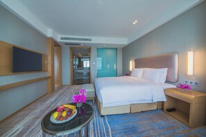 Een hotelkamer met een groot bed, een flatscreen televisie aan de muur, een klein rond tafeltje met fruit en bloemen, en een badkamer zichtbaar door een geopende deur.