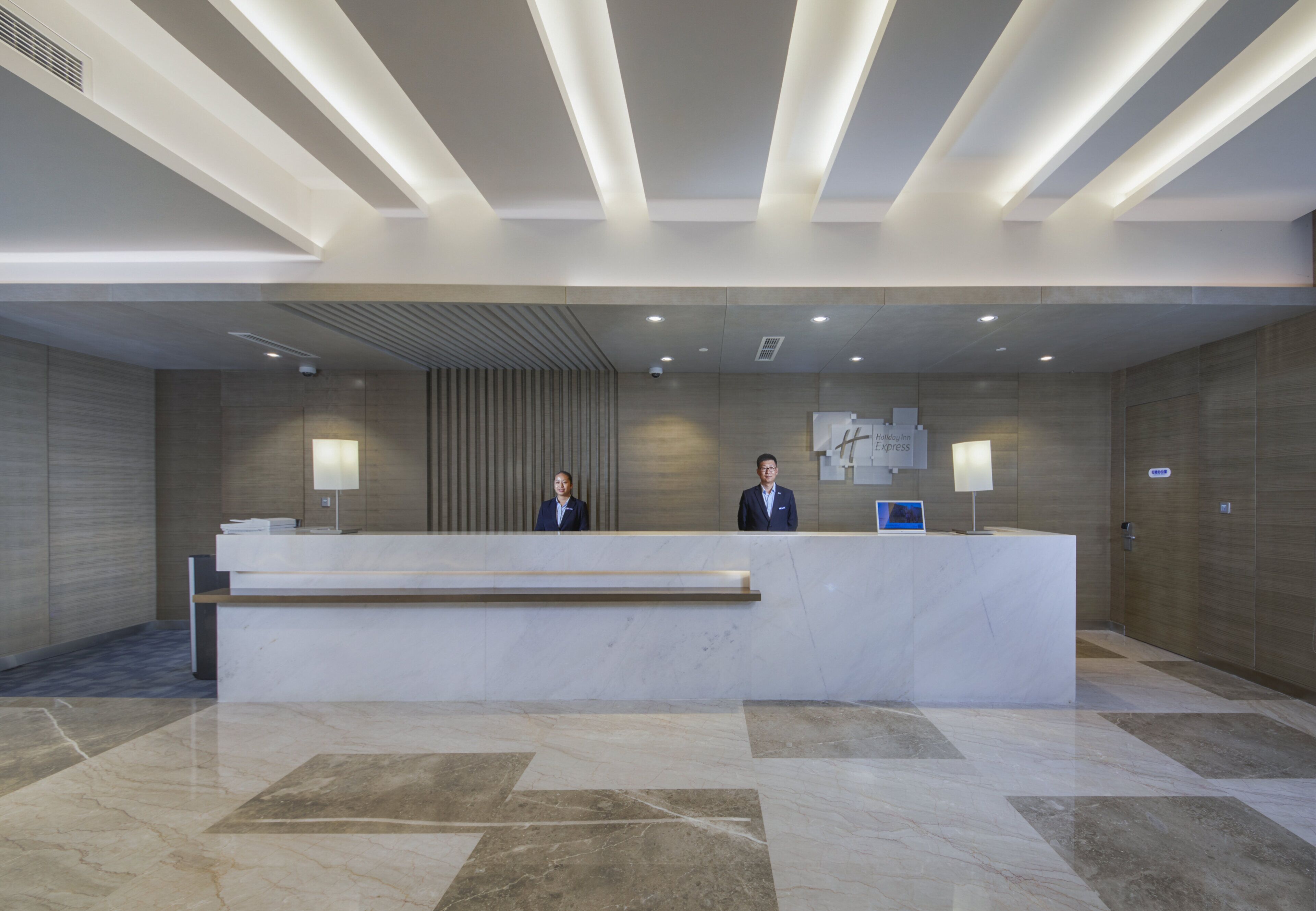 Un comptoir d’accueil d’hôtel avec deux employés, un design moderne et un écran numérique.