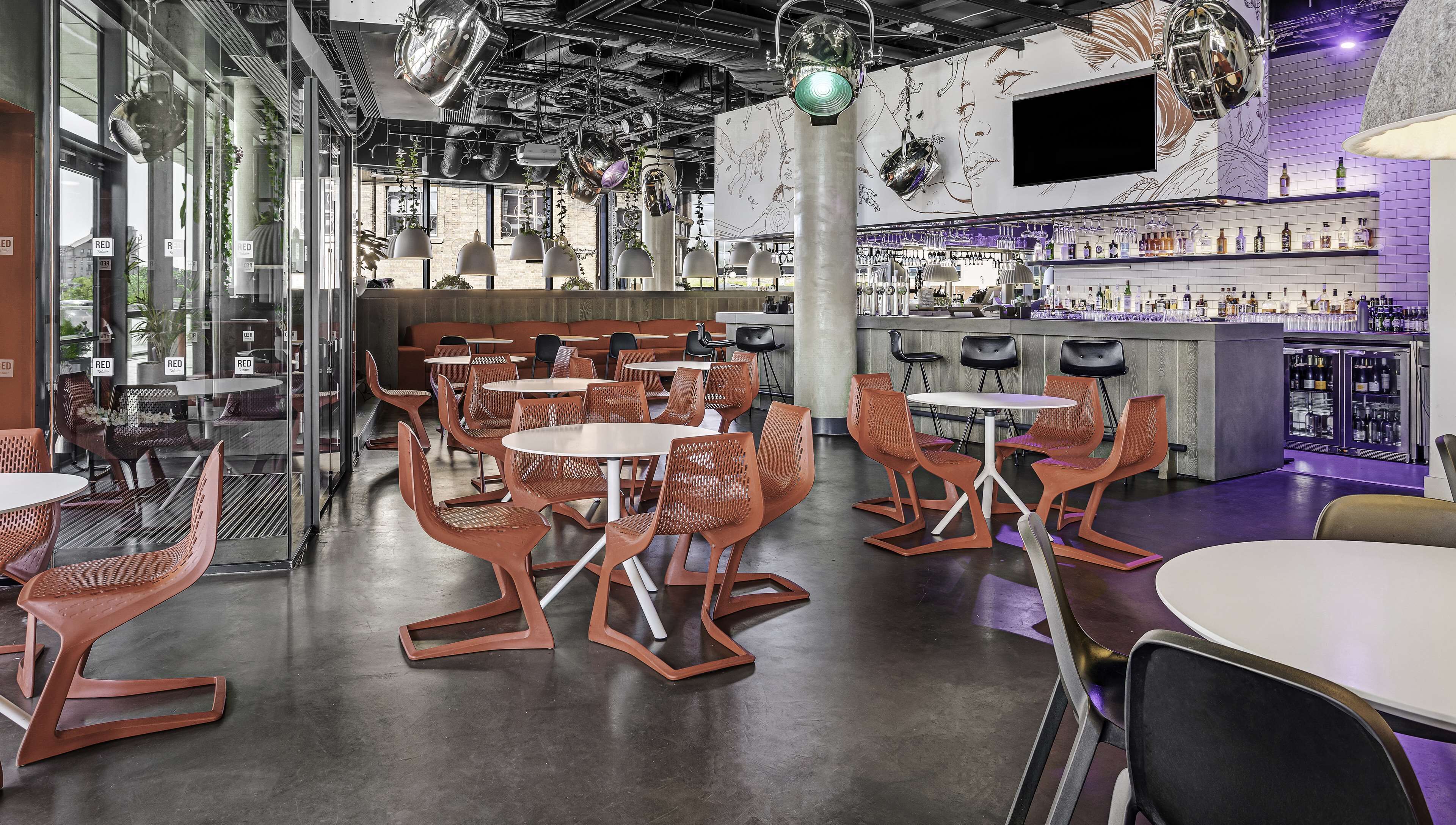 A modern bar area with round tables and orange chairs, a bar counter with various bottles, and a large mural on the wall.