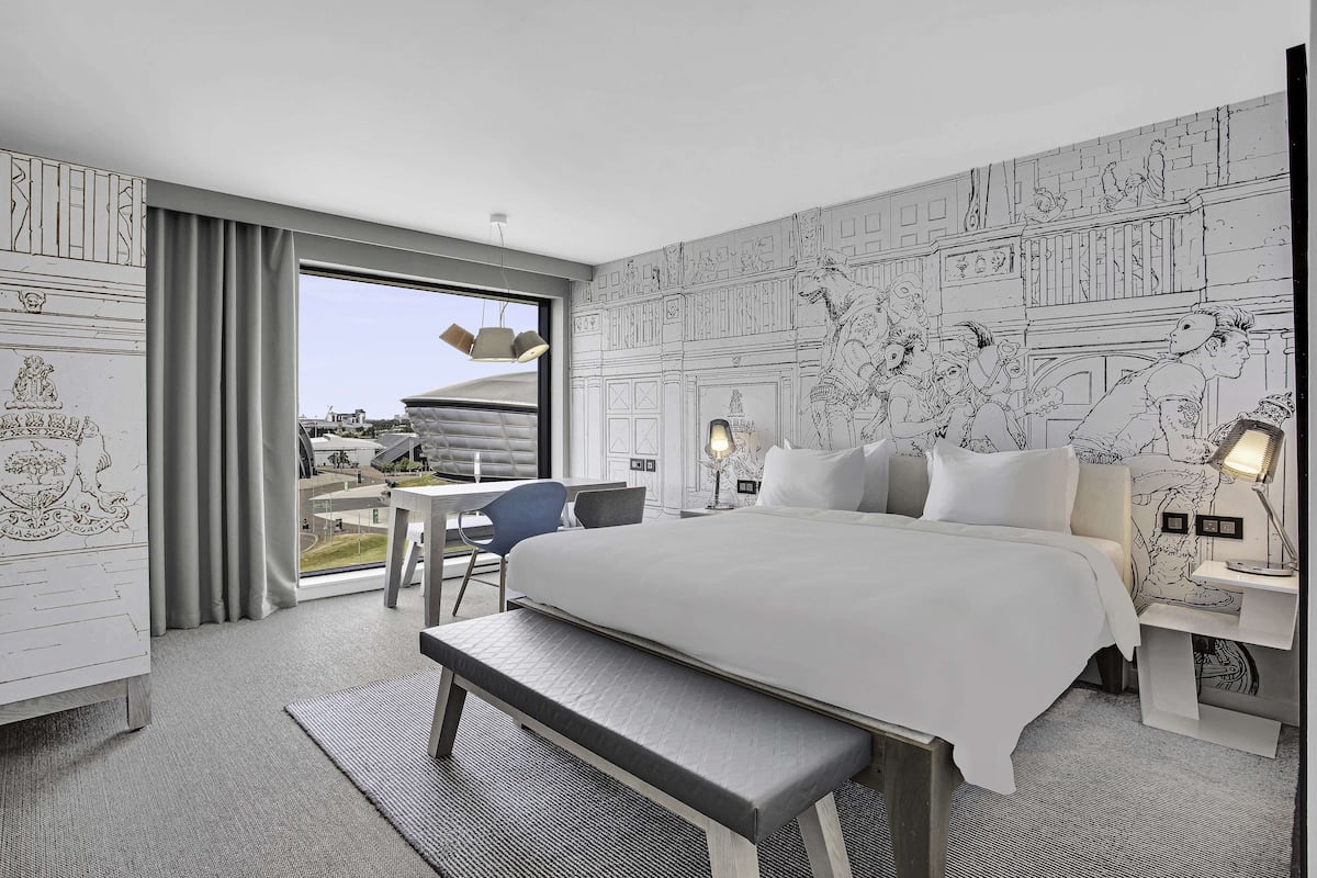 Ein modernes Hotelzimmer mit einem großen Bett, einer Sitzbank, einem Schreibtisch und Blick auf die Stadt durch große Fenster.