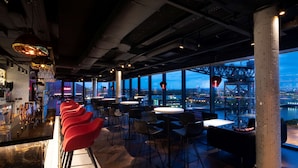 Zona de bar moderna con taburetes rojos, mesas redondas y vistas de la ciudad a través de amplios ventanales.