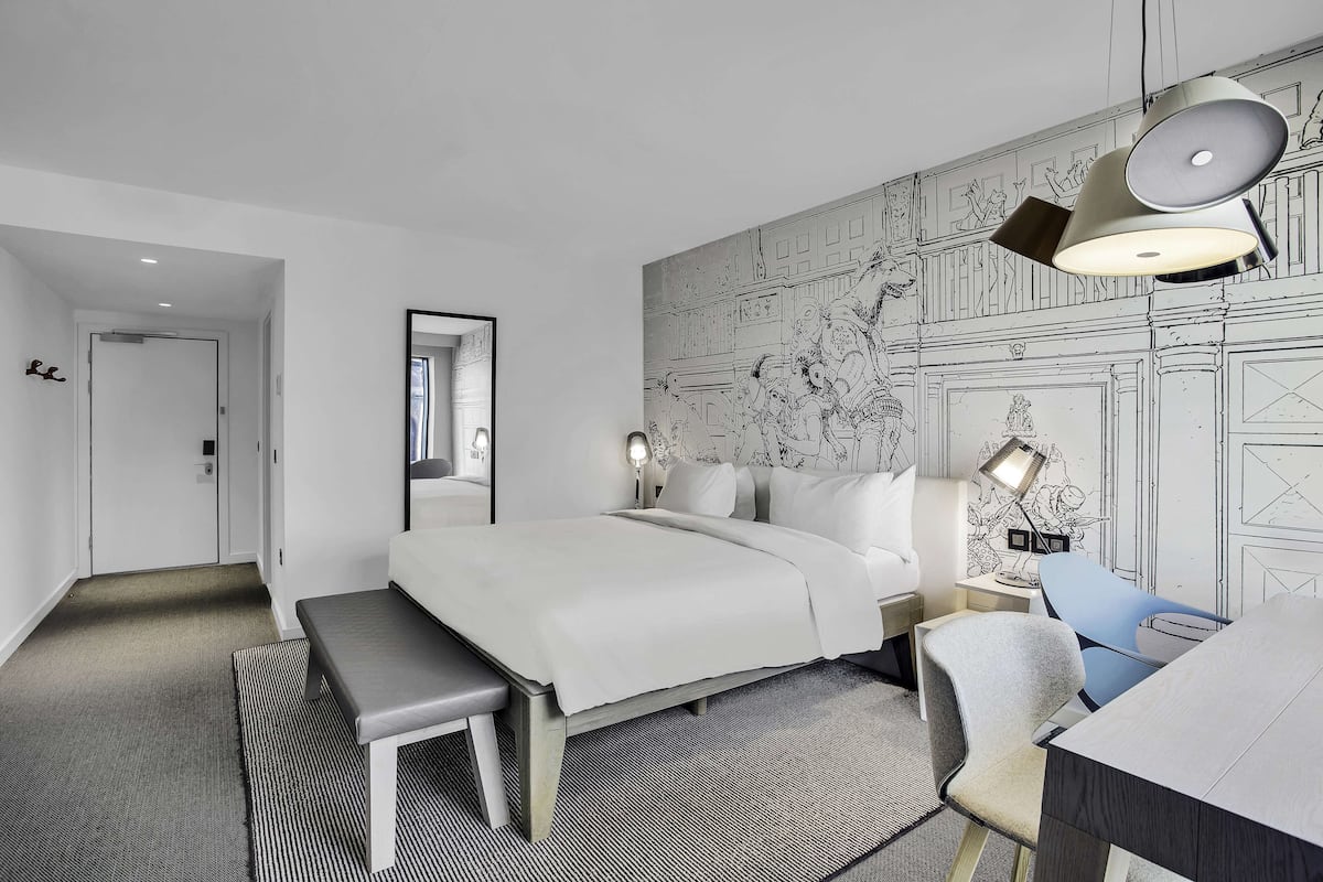 Ein modernes Hotelzimmer mit einem großen Bett, einem Schreibtisch und einem Sessel. An der Wand ist ein Wandbild angebracht, und eine Pendelleuchte hängt von der Decke.