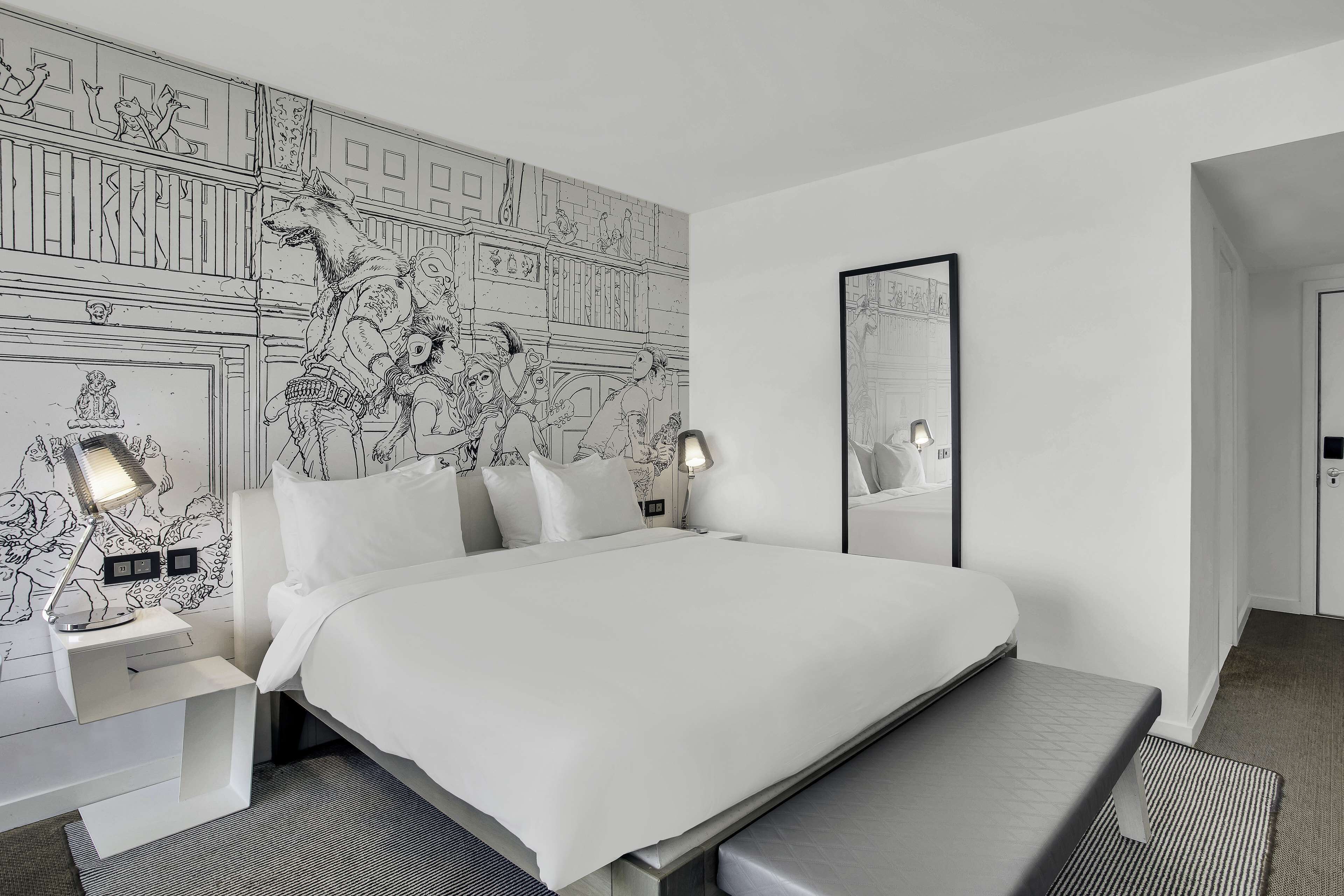 A modern hotel room with a large bed, bedside tables with lamps, a bench, and a mural on the wall.