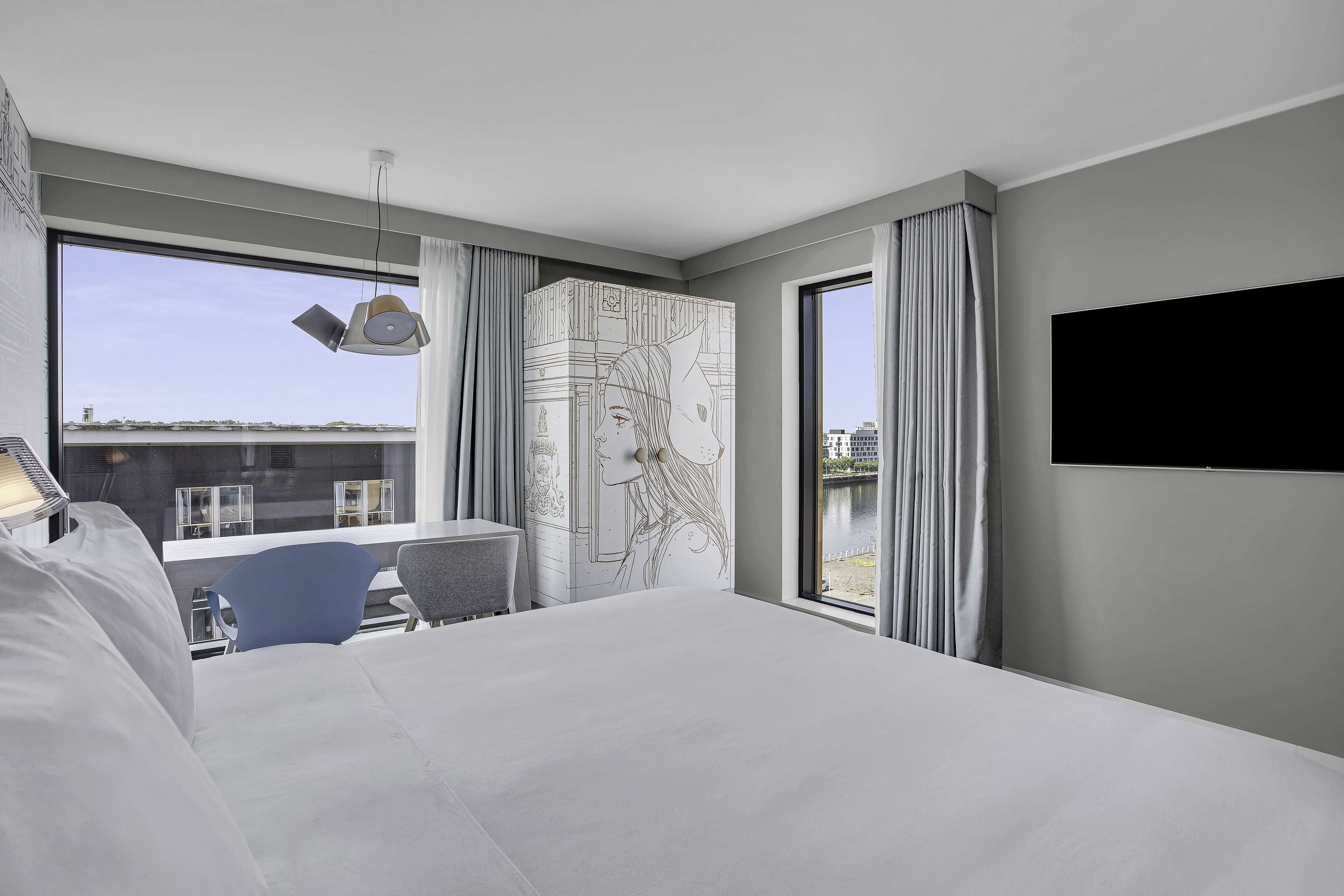 A modern hotel room with a large bed, a desk, and a chair. There is a large window with curtains, a flat-screen TV mounted on the wall, and a map on the wall.