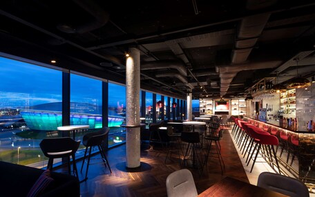A modern bar area with high tables and red bar stools, large windows offering a view of a cityscape, and a prominent pillar in the center.