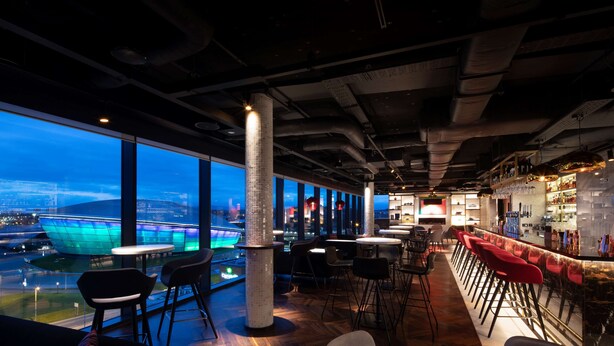 Zona de bar moderna con mesas altas y taburetes rojos, ventanas amplias que ofrecen vistas de un paisaje urbano y un pilar destacado en el centro.