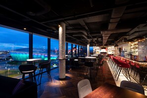 A modern bar area with high tables and red bar stools, large windows offering a view of a cityscape, and a prominent pillar in the center. - Radisson RED Glasgow (Glasgow)