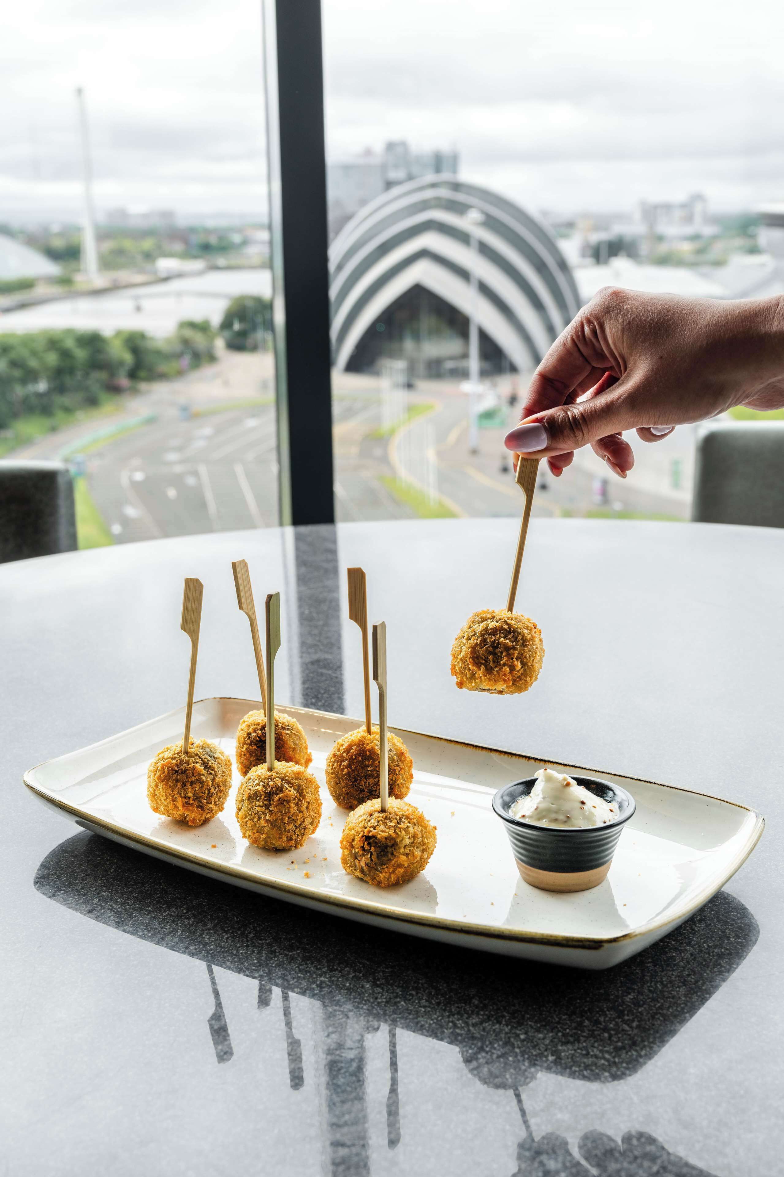 A hand holding a croquette with a toothpick, a plate with croquettes, and a small bowl of sauce on a table with a view of a cityscape.