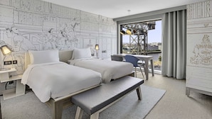 Habitación de hotel con dos camas, un banco, un escritorio y vistas a un paisaje urbano.