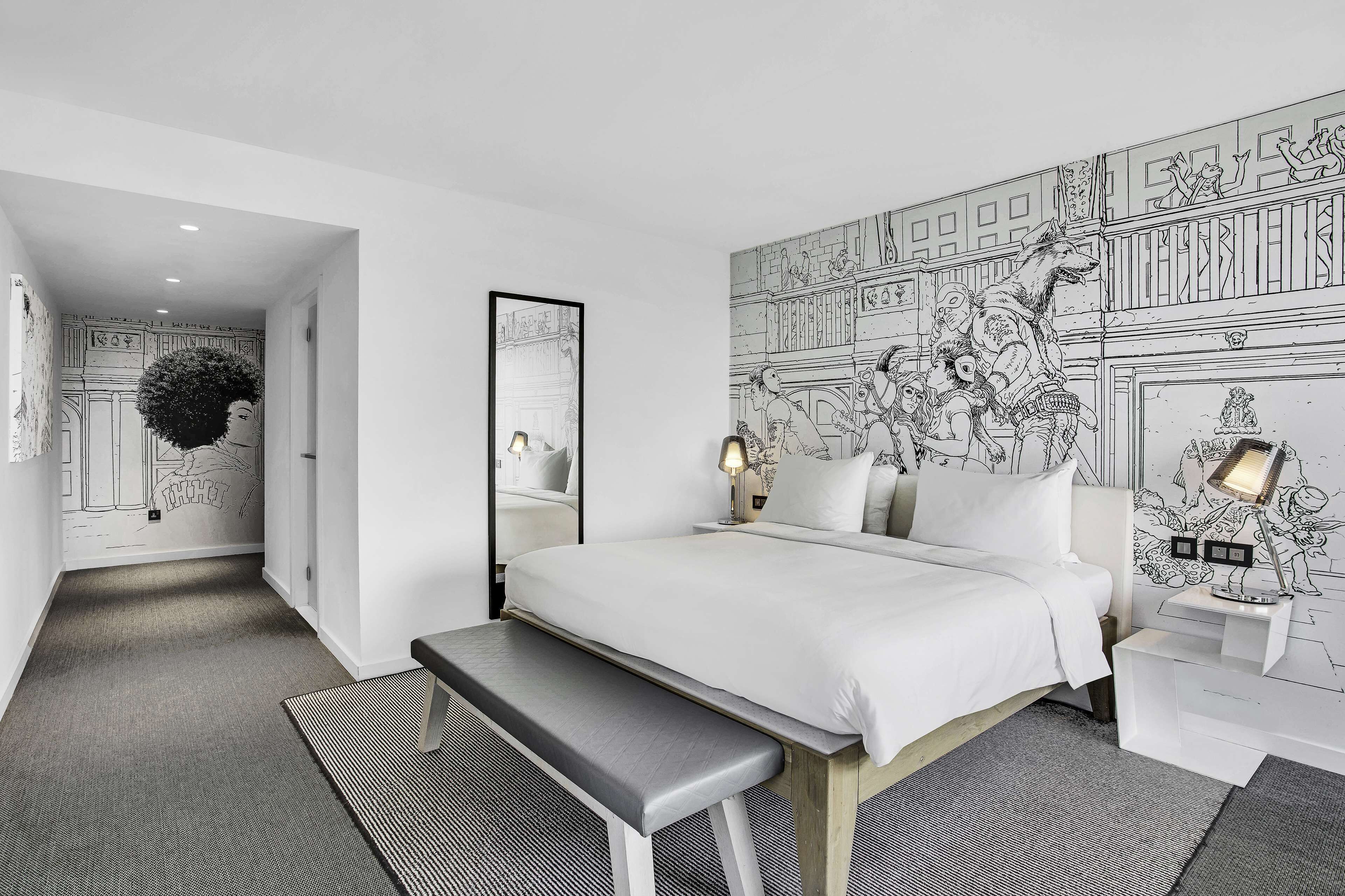 Una habitación de hotel moderna con una cama grande, un banco, una mesita de noche con lámparas y un mural en la pared.