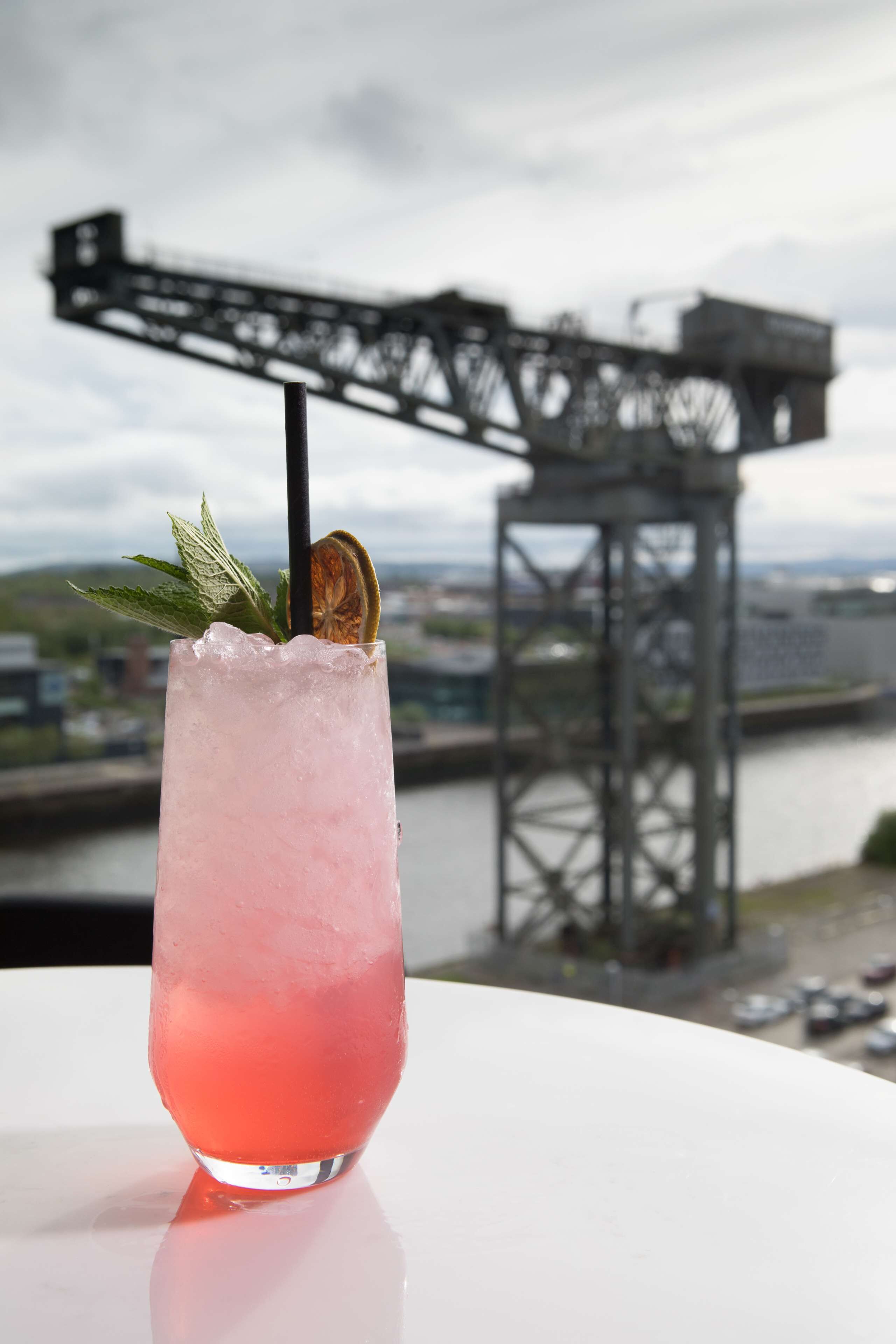 Um cocktail rosa com hortelã e uma fatia de limão, colocado sobre uma superfície branca com um rio e uma ponte ao fundo.