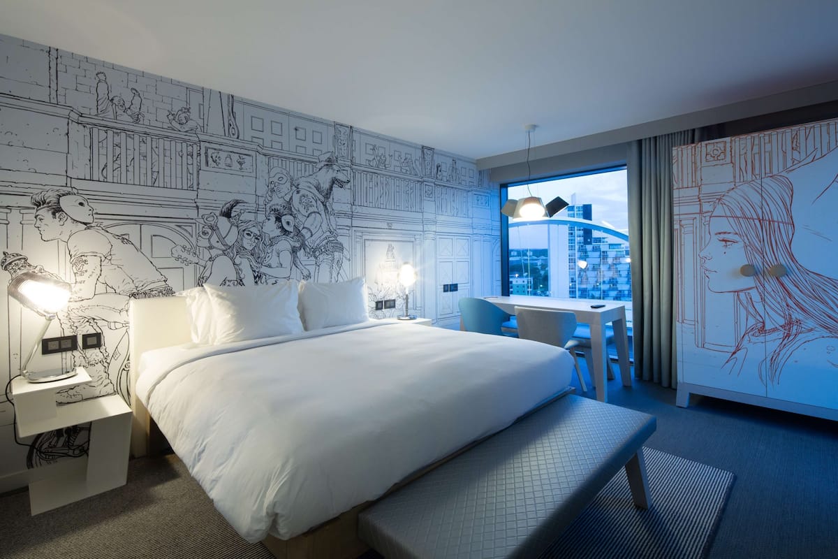 Ein Hotelzimmer mit einem großen Bett, einem Schreibtisch und Blick auf die Stadt.
