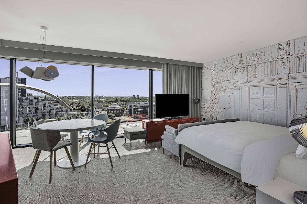 Ein modernes Hotelzimmer mit einem großen Bett, einem Schreibtisch mit Flachbildfernseher, einer Essecke mit rundem Tisch und Stühlen sowie Blick auf die Stadt durch große Fenster.