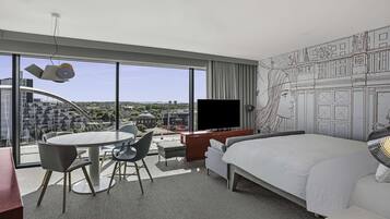 Una habitación de hotel moderna con una cama grande, un escritorio con televisor de pantalla plana, un rincón de comedor con mesa y sillas redondas, y vistas del paisaje urbano a través de amplias ventanas.