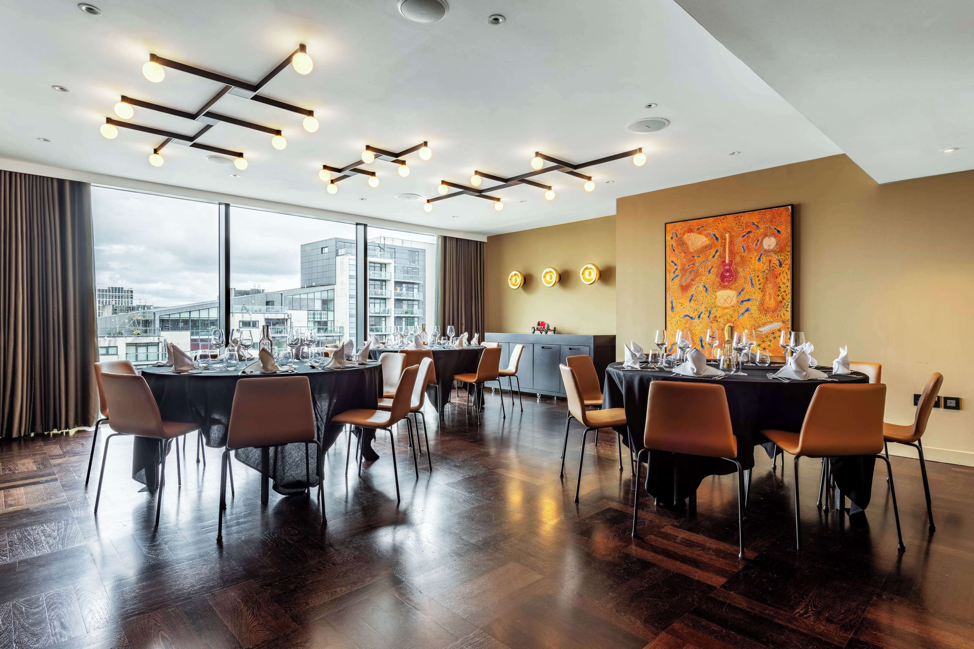 Un comedor moderno con varias mesas dispuestas para una comida, amplias ventanas con vistas a la ciudad e iluminación contemporánea.