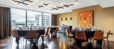 Un comedor moderno con varias mesas dispuestas para una comida, amplias ventanas con vistas a la ciudad e iluminación contemporánea.